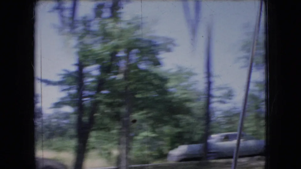 1966 Ohio 8mm frame with motion-blurred trees, pale sky, roadside greenery, and a partly visible vehicle passing or parked beyond the foliage.
