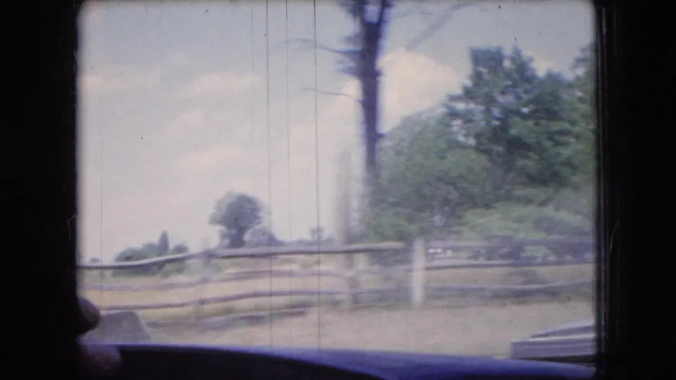 1966 Ohio 8mm home-movie frame from inside a moving car, showing wooden fencing, roadside trees, grass, pale sky, window framing, and a dark dashboard edge.