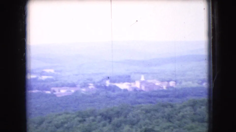 1966 Ohio home-movie frame looking across wooded hills toward a hazy distant town or settlement, with bright exposure, window framing, and visible film scratches.