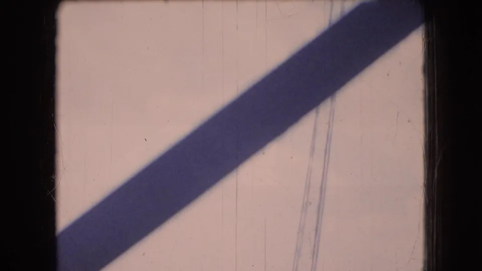 Final 8mm frame from the Ohio tower sequence showing a strong diagonal shadow across a pale surface, with faint scratches and vertical marks in the film image.