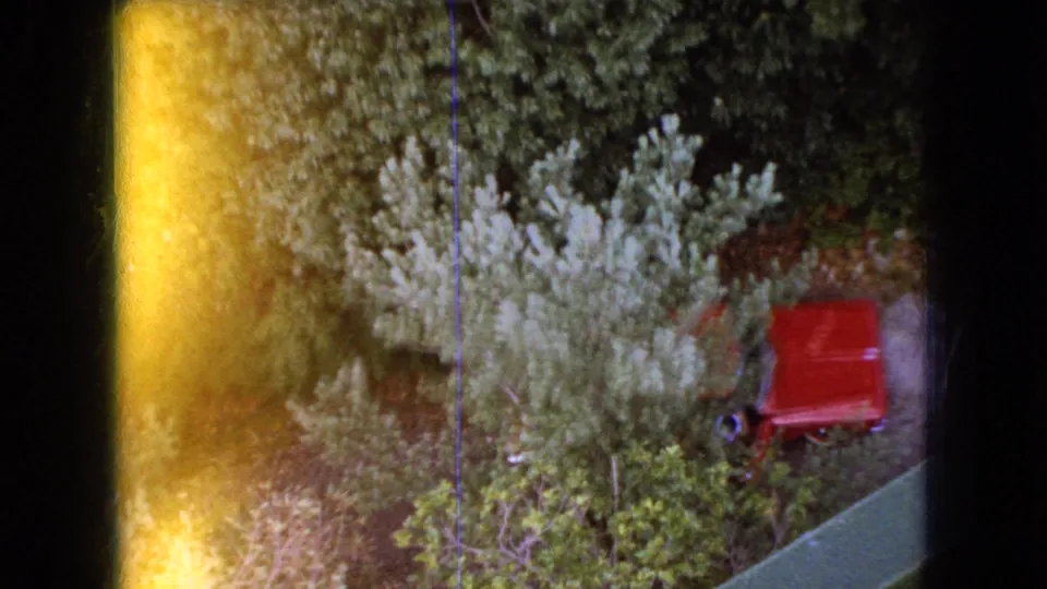 1966 Ohio 8mm downward view across treetops from a framed elevated position, with a red object among foliage and a bright yellow-orange film flare at the left.