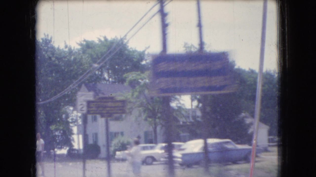 Street-side view with parked cars, houses or storefronts, utility poles, and a blurred roadside sign.
