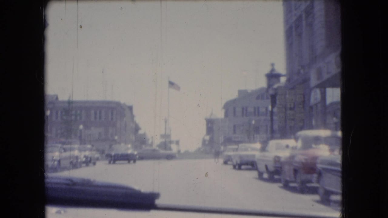 1966 Ohio street scene filmed from inside a moving car, with parked classic cars, two-story buildings, a flagpole, and traffic visible through the windshield.