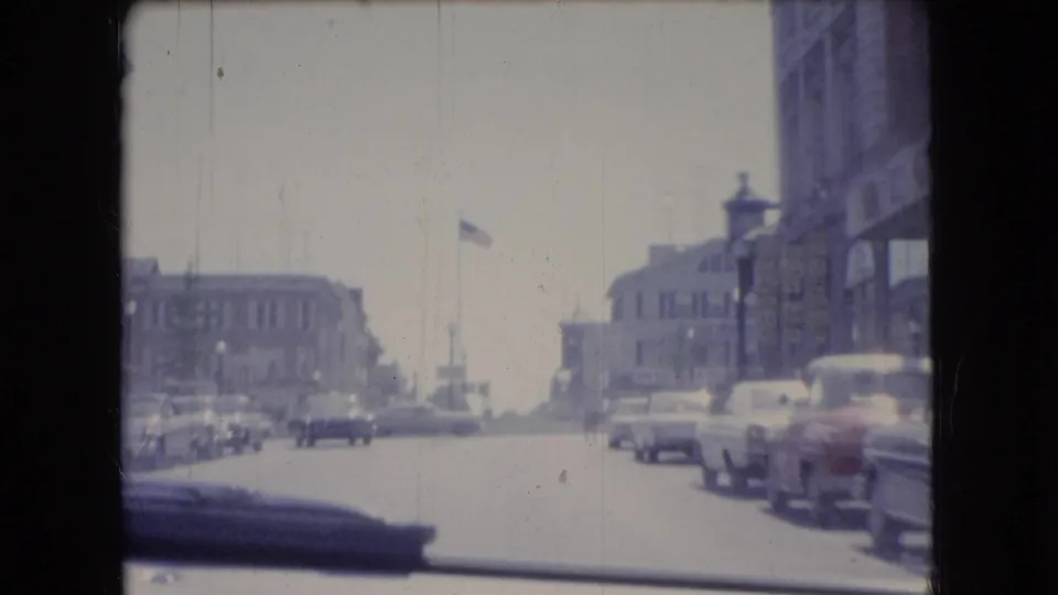 1966 Ohio street scene filmed from inside a moving car, with parked classic cars, two-story buildings, a flagpole, and traffic visible through the windshield.