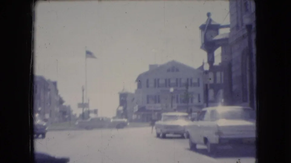 1966 Ohio home-movie frame showing a downtown intersection with a light-colored station wagon, storefront buildings, parked cars, and an American flag above the street.