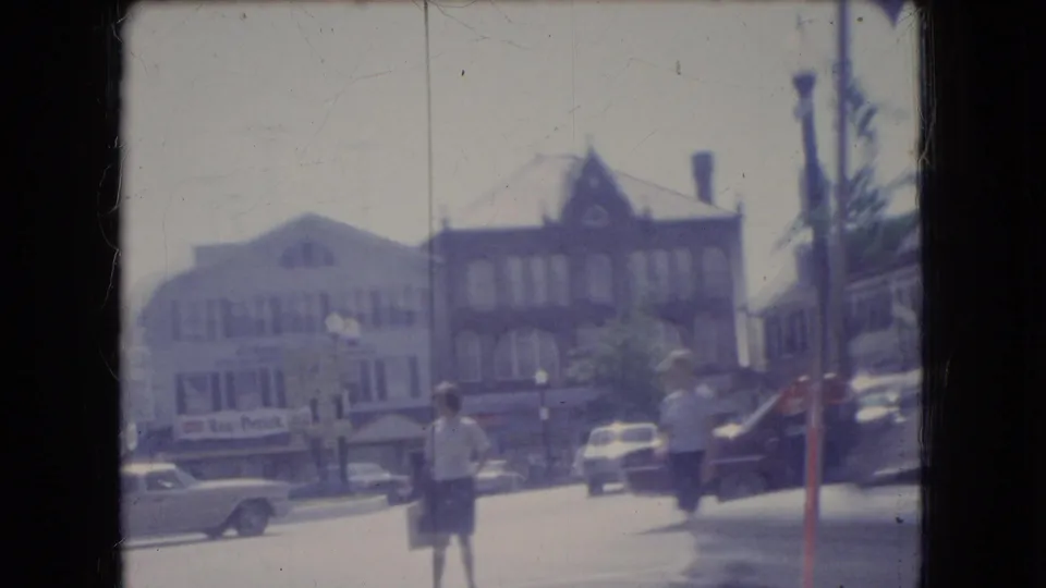 1966 Ohio street corner with brick commercial buildings, storefront signs, parked classic cars, and a pedestrian in a light shirt and dark shorts crossing or standing near the roadway.