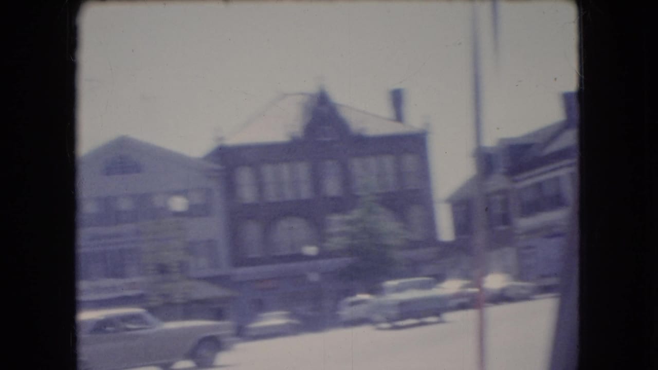 1966 Ohio 8mm home-movie frame showing a soft, motion-blurred view of brick storefronts, parked sedans, and a town street from a moving vehicle.