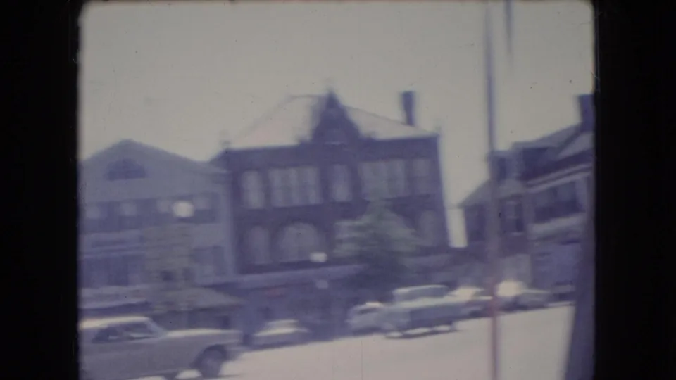 1966 Ohio 8mm home-movie frame showing a soft, motion-blurred view of brick storefronts, parked sedans, and a town street from a moving vehicle.
