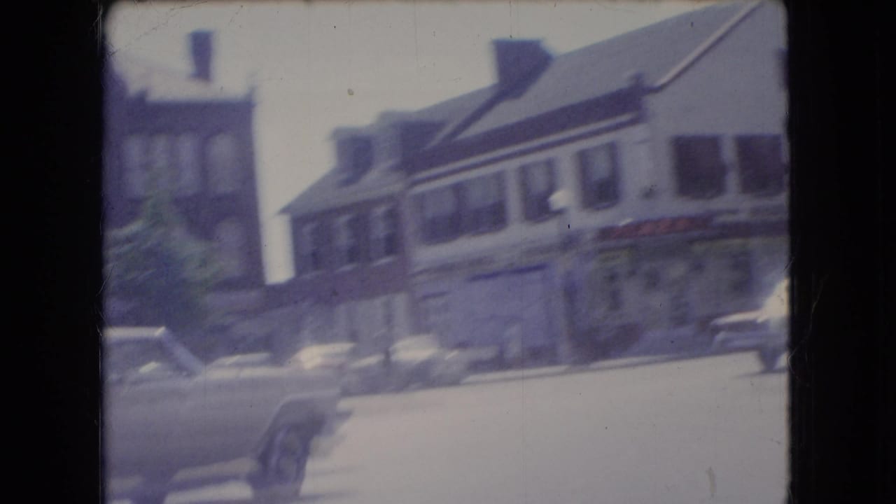 1966 Ohio street view from a moving car, with storefronts, parked vehicles, and a briefly visible person walking or standing near the curb.
