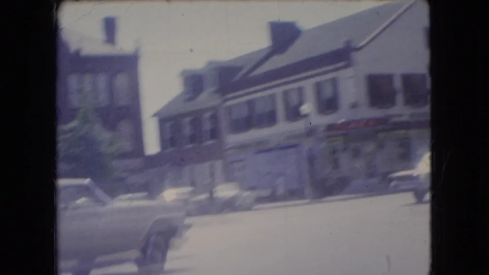 1966 Ohio street view from a moving car, with storefronts, parked vehicles, and a briefly visible person walking or standing near the curb.