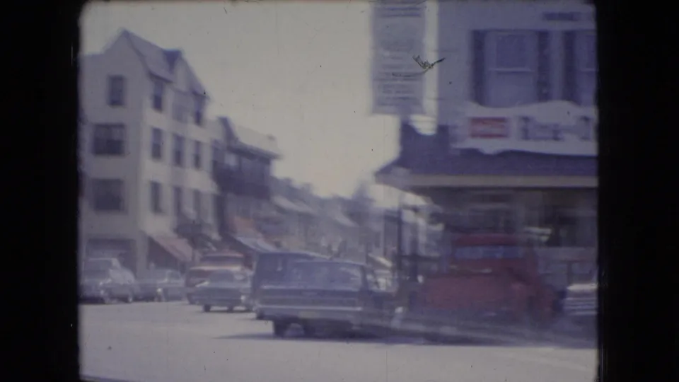Soft 8mm frame of an Ohio town street in 1966, showing multistory buildings, storefront signage, parked cars, and a station wagon from a moving-vehicle viewpoint.