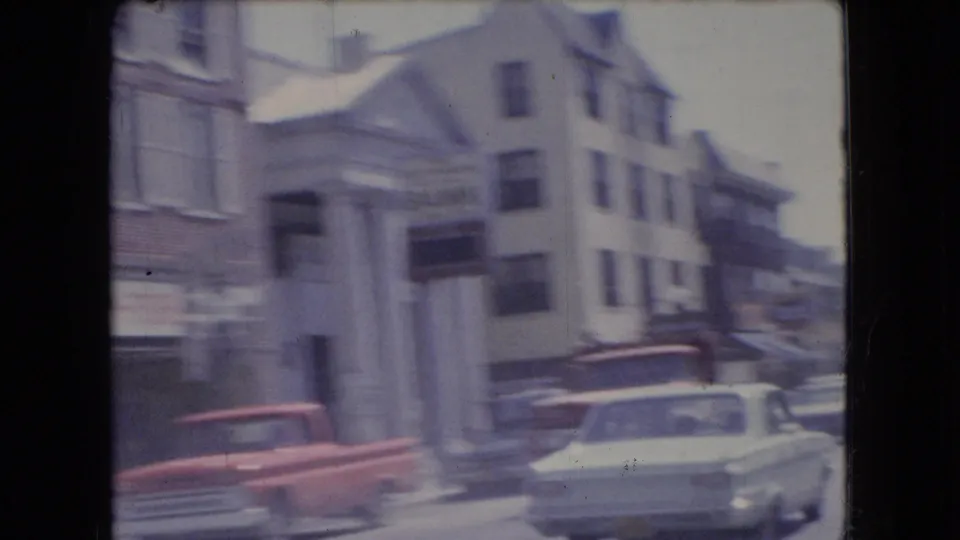 1966 Ohio moving-car frame showing a town intersection with storefronts, houses, a red pickup or utility vehicle, and a white sedan under faded 8mm color.