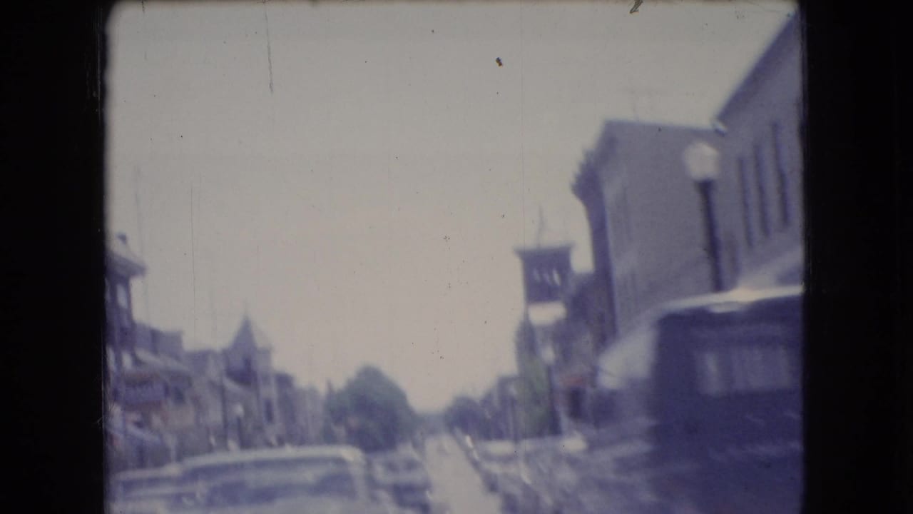 Soft 8mm view of a downtown Ohio street in 1966, filmed from a moving vehicle with low-rise buildings, parked cars, and a dark van ahead.