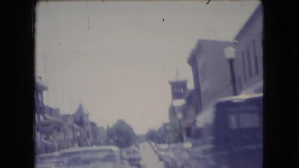 Soft 8mm view of a downtown Ohio street in 1966, filmed from a moving vehicle with low-rise buildings, parked cars, and a dark van ahead.