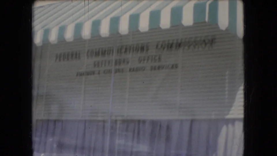 Blurred 8mm frame of a striped-awning storefront or office facade in Ohio, with a visible Federal Communications Commission sign on the wall.