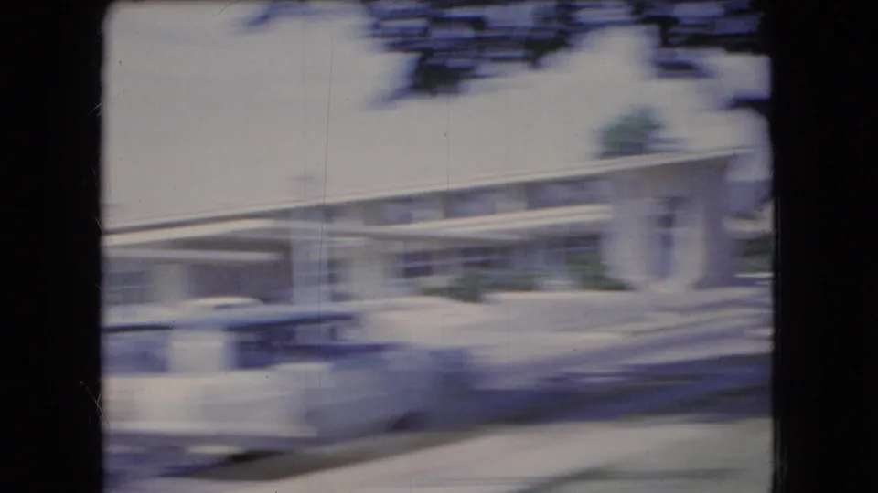 Moving-car 8mm view of a sunlit Ohio streetscape with classic cars, a low commercial building, canopy-like roofline, and roadside blur.