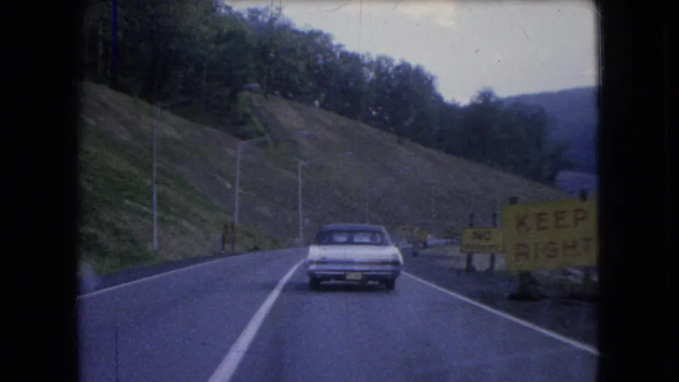 Clearer 1960s road view from a moving vehicle, showing a vintage car ahead on a two-lane road beside wooded hills and construction-related roadside signs.