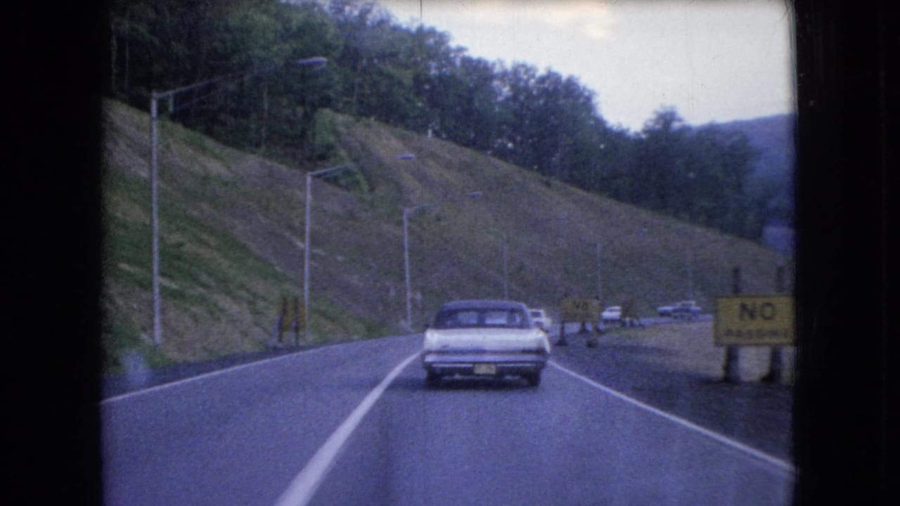 1966 Ohio home-movie frame of a classic sedan traveling along a winding road bordered by a steep grassy embankment, trees, roadside poles, and a yellow no-passing sign.