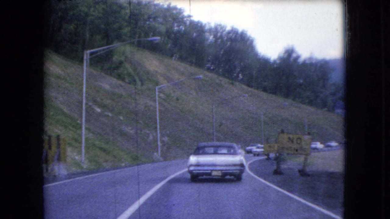Car ahead follows a bend in the road with hillside, trees, traffic, and a roadside sign.