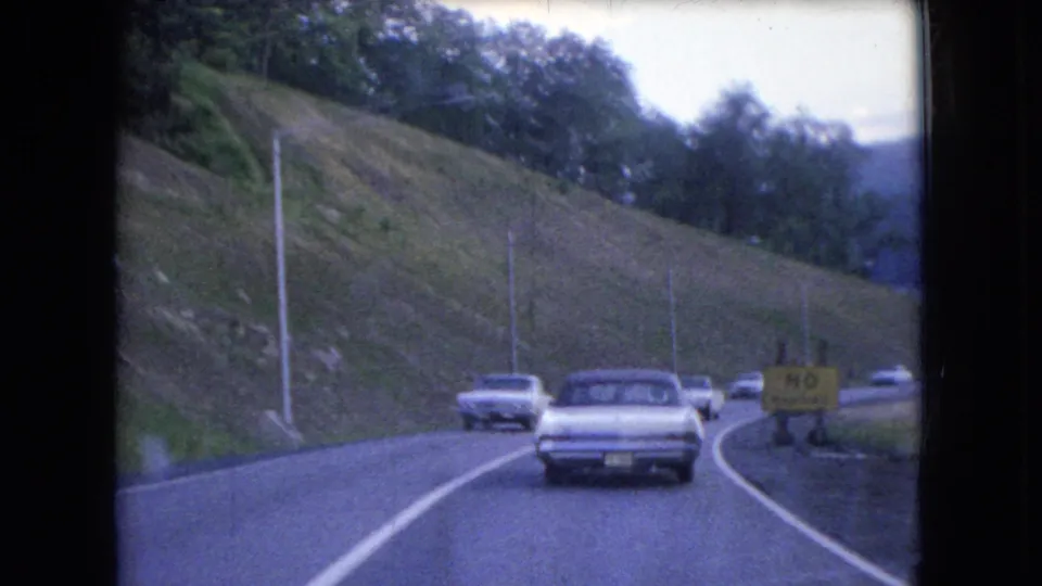 The frame captures 1966 Ohio driving footage with a sedan ahead, an oncoming car, a winding roadway, a grassy embankment, roadside poles, trees, and a yellow no-passing sign.