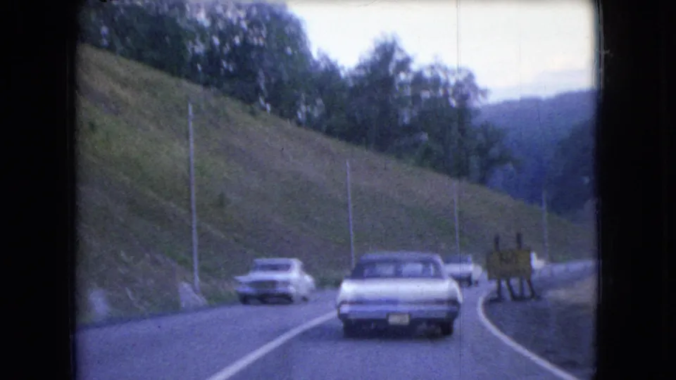 A later 1966 Ohio home-movie frame shows the car ahead nearing a road bend while another vehicle passes in the opposite lane, with wooded hills and roadside signage visible.