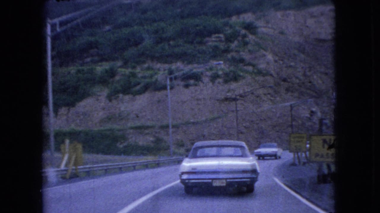 A white 1960s sedan drives ahead on a curved Ohio hillside road in 1966, with guardrails, utility poles, and roadside signs visible in the 8mm frame.