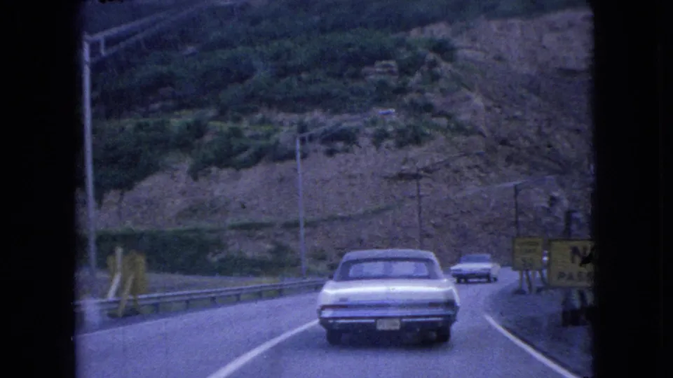 A white 1960s sedan drives ahead on a curved Ohio hillside road in 1966, with guardrails, utility poles, and roadside signs visible in the 8mm frame.