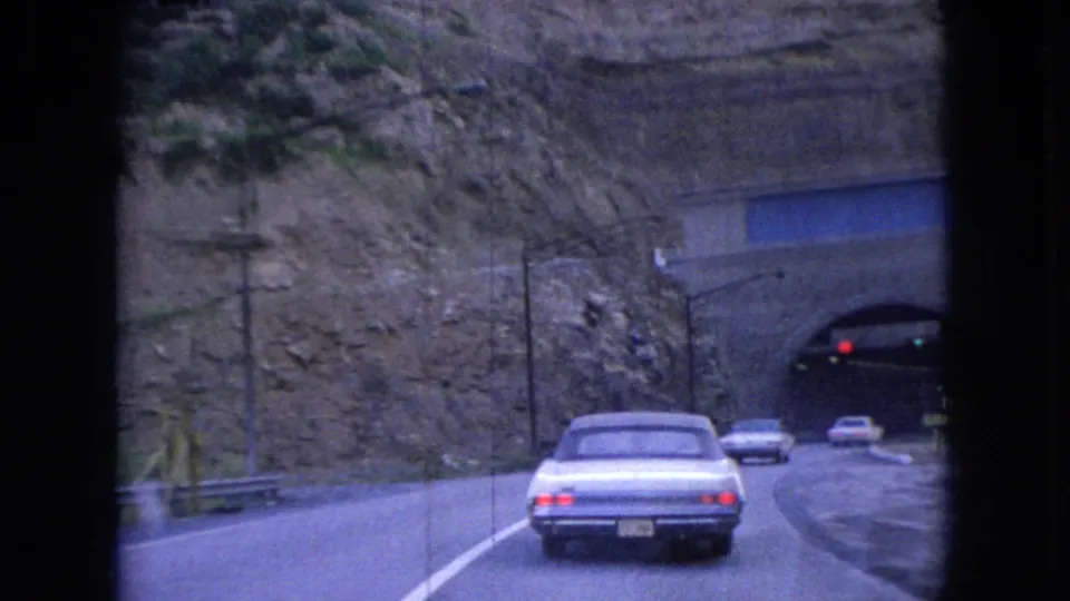 A white sedan nears a tunnel entrance on a curving Ohio road in 1966, with traffic lights, guardrails, utility poles, and a steep rock face in view.