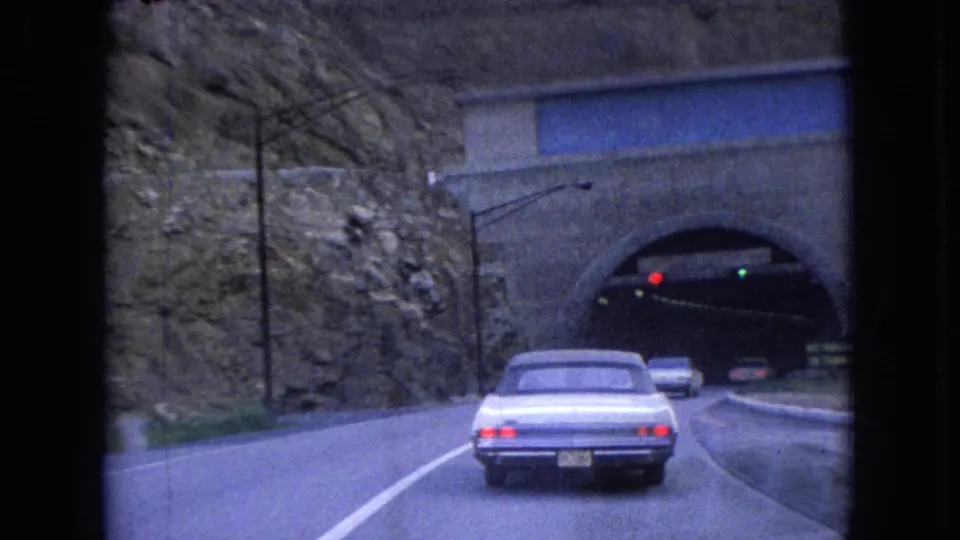 The white sedan approaches the tunnel portal on the 1966 Ohio roadway, with red and green traffic lights, other cars, guardrails, and the rocky hillside around the entrance.