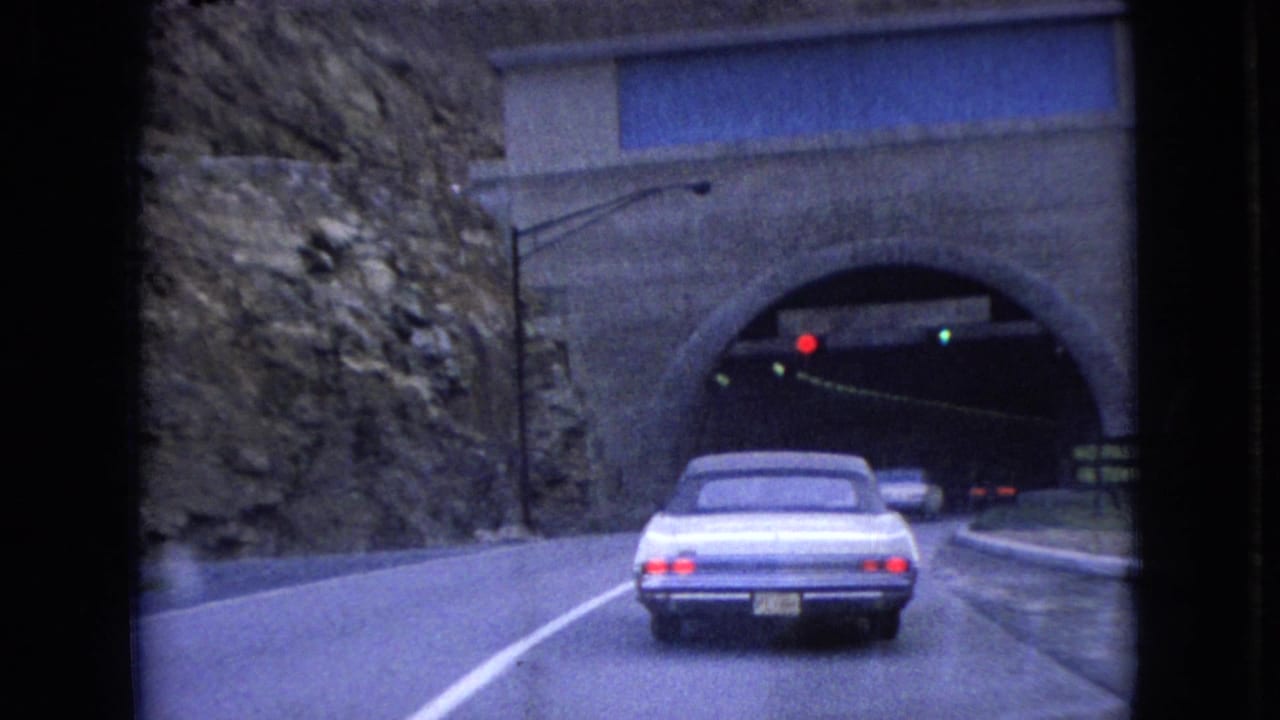In 1966 Ohio home-movie footage, a light-colored classic sedan drives toward a stone-arched tunnel with red and green traffic signals above the entrance.