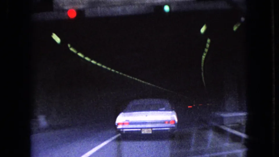 The classic sedan is partly inside the tunnel, its rear visible below dim ceiling lights and signal glow as the camera follows from the roadway behind.
