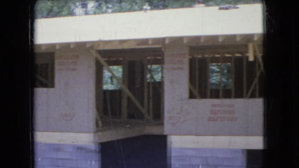 The 1966 Ohio home-movie frame shows a partially built house with pale wall sheathing, open framed bays, diagonal supports, roof-edge framing, and a concrete-block foundation.