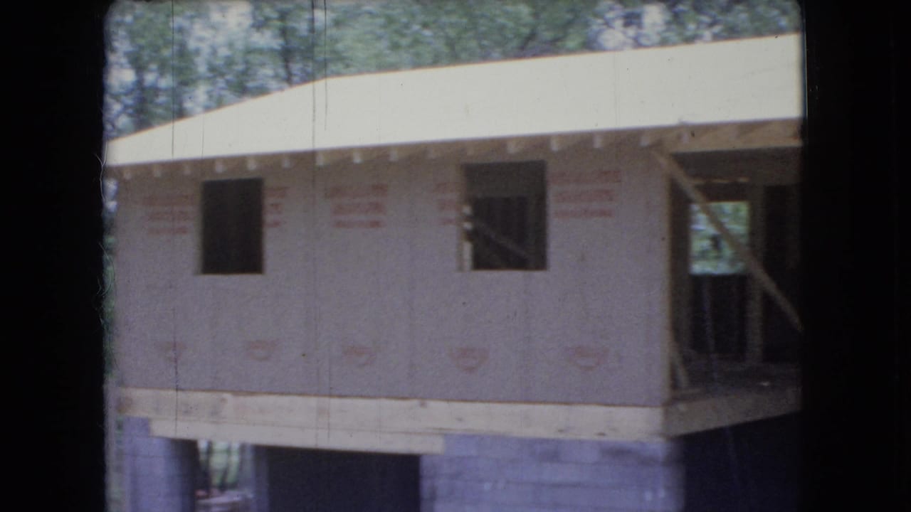 1966 Ohio 8mm frame showing an unfinished residential building with plywood sheathing, open window cutouts, exposed entry framing, concrete block foundation, and trees behind it.