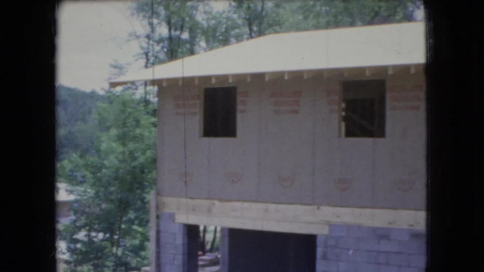 1966 Ohio home-movie frame of a partially built house on a block foundation, with light wall sheathing, dark open window spaces, roof overhang, and surrounding green trees.