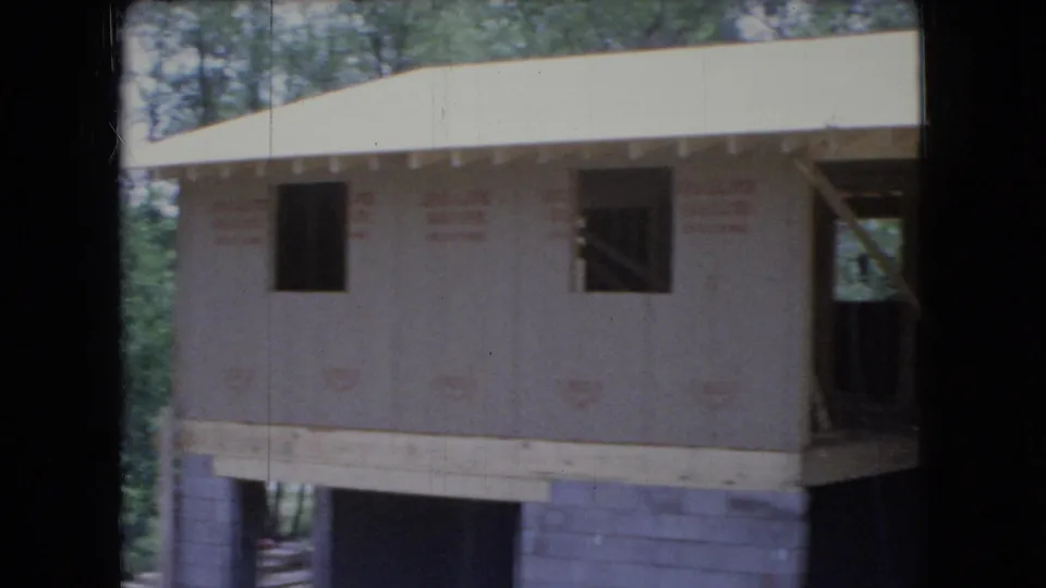 1966 Ohio 8mm frame showing a closer exterior view of a residential construction project, including plywood sheathing, open window cutouts, exposed lumber, concrete block support, and trees at the lot edge.