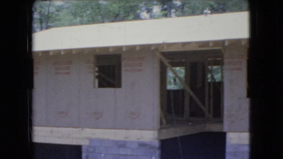1966 Ohio home-movie frame showing the corner of an unfinished house with open windows, exposed entry or porch framing, plywood sheathing, a concrete block base, and leafy trees behind the structure.