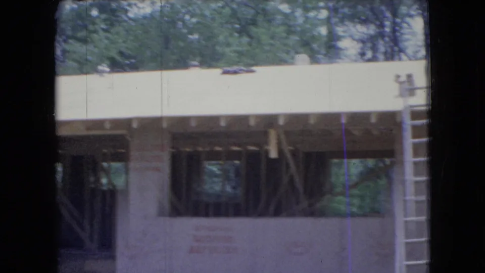 1966 Ohio 8mm frame showing the framed side of a rural house under construction, with sheathing, open wall bays, trees, and a ladder near the right edge.