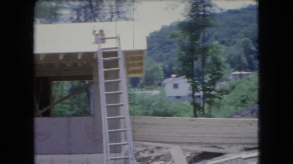 1966 Ohio 8mm frame showing a ladder next to the partially built country house, with loose lumber, a small distant building, and a wooded hillside in the background.