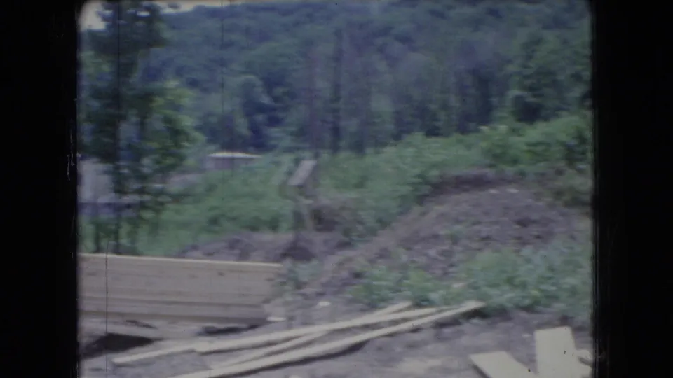 1966 Ohio 8mm frame looking away from the house toward the rural construction site, showing scattered lumber, disturbed soil, dense trees, and a small distant building.
