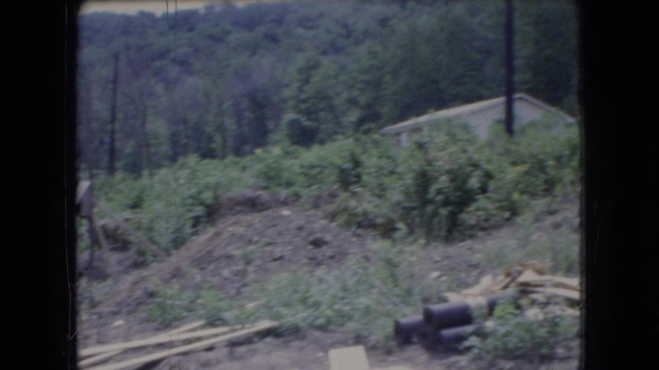 1966 Ohio 8mm frame showing a wooded hillside near a house construction site, with scattered materials, earthwork, and a distant structure partly obscured by soft focus.