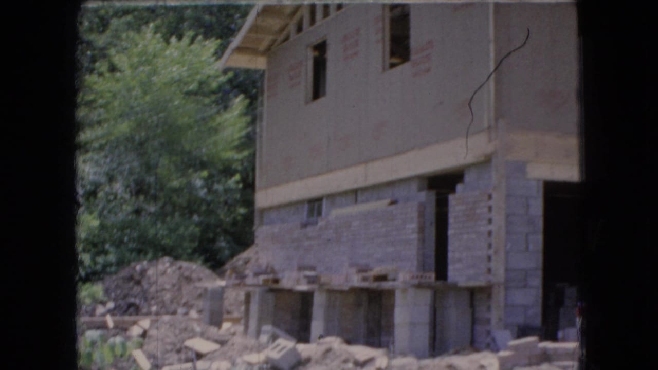 Unfinished house with concrete block lower walls and framed upper story beside trees.