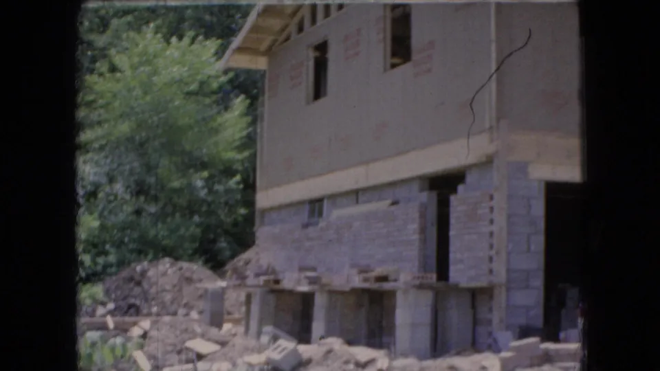 1966 Ohio home-movie frame of an unfinished house in a wooded area, showing concrete block walls, open window spaces, exposed upper framing, rubble, and construction materials.