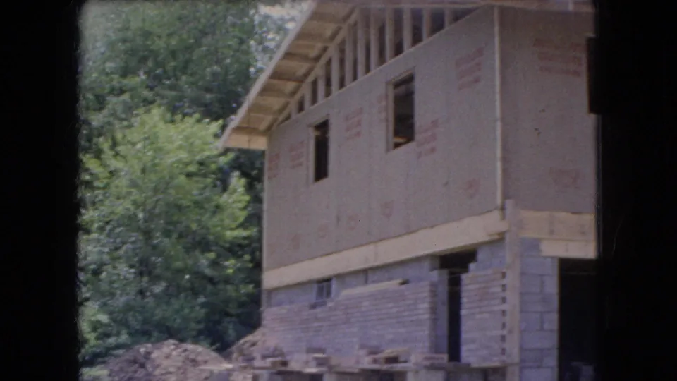 1966 Ohio 8mm frame showing the side of a house under construction, with an unfinished upper story, concrete block lower level, wooded background, and dirt piles around the work area.