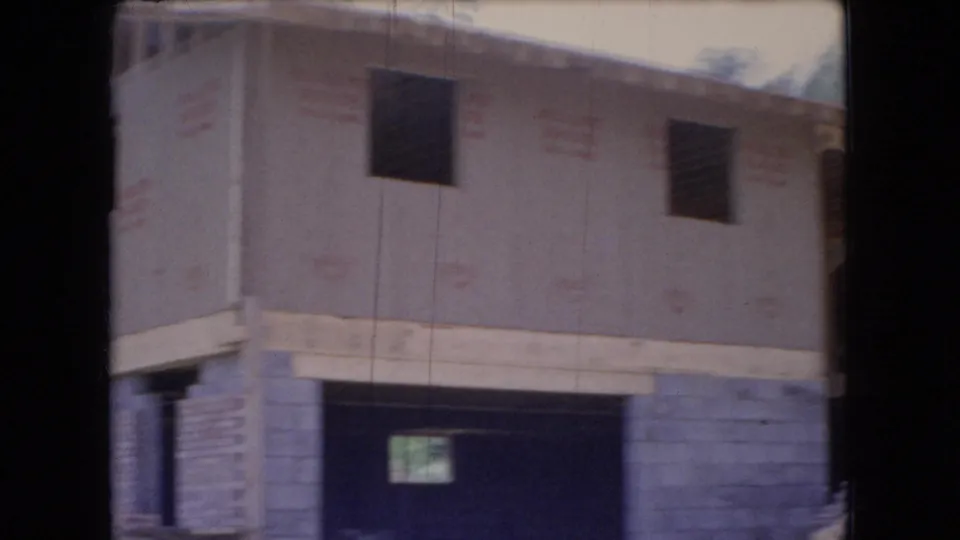 1966 Ohio home-movie frame showing a closer view of the unfinished house, including open window cutouts, pale exterior sheathing, concrete block walls, a dark lower opening, and wooded surroundings.