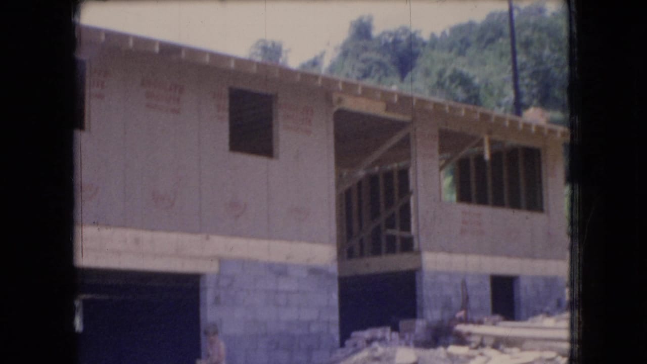 1966 Ohio home-movie frame of a new house being built, showing sheathed exterior walls, exposed framing, a concrete block base, loose boards, dirt piles, and wooded background.
