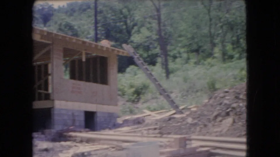 1966 Ohio 8mm frame showing the side of a house under construction beside a wooded slope, with a ladder, exposed framing, sheathing, dirt piles, and scattered lumber.