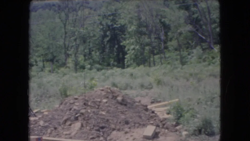1966 Ohio home-movie frame looking across the construction site toward trees, with a dirt pile, scattered lumber, grass, and the edge of the unfinished house at left.