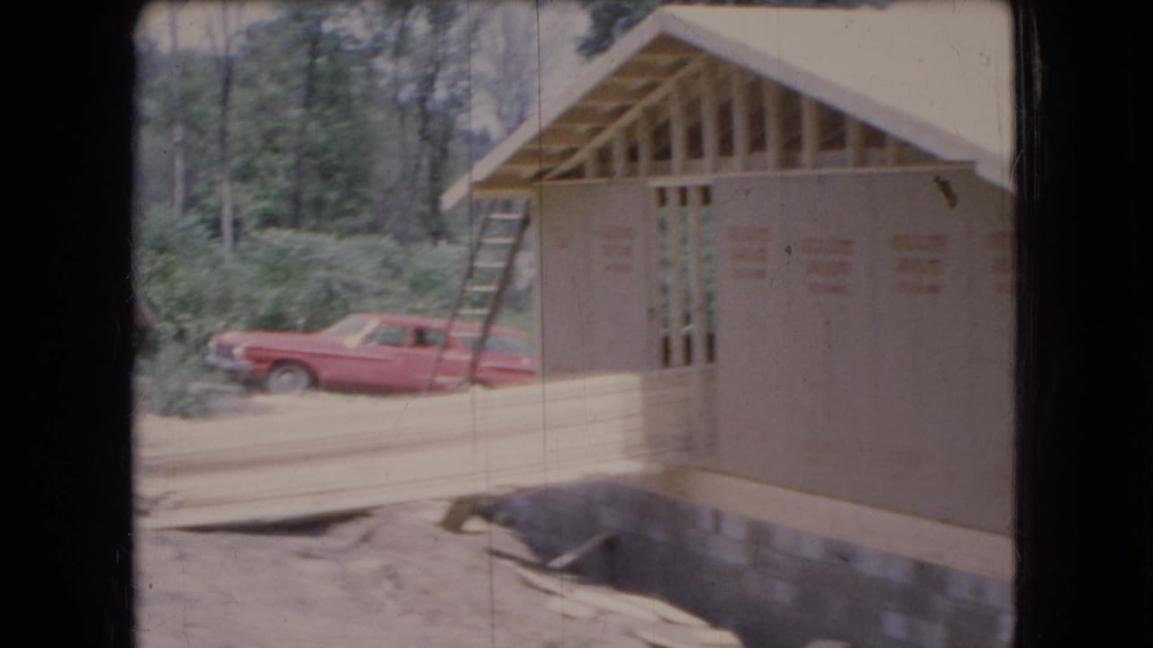 Ohio 1966 8mm frame showing an unfinished house under construction with plywood sheathing, exposed roof framing, a ladder, wooded background, and a red station wagon parked to the side.