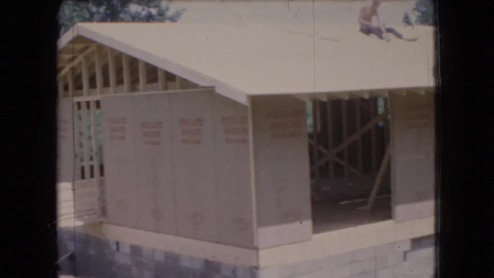 Ohio 1966 home-movie frame of a partially built house with plywood wall panels, open doorway framing, roof framing, concrete block foundation, and wooded surroundings.