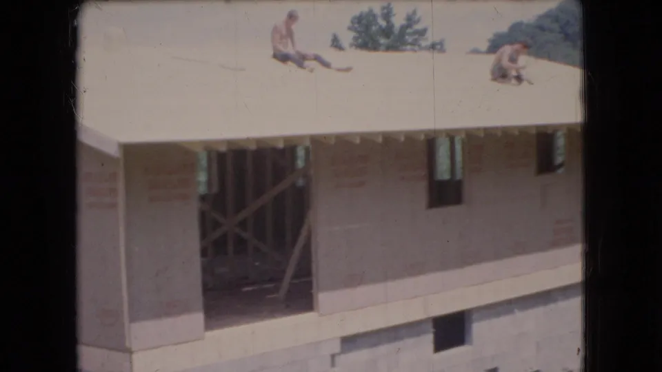Ohio 1966 8mm frame showing two men on the roof of an unfinished suburban house, with exposed framing, sheathed walls, a dark doorway opening, and concrete block foundation visible below.